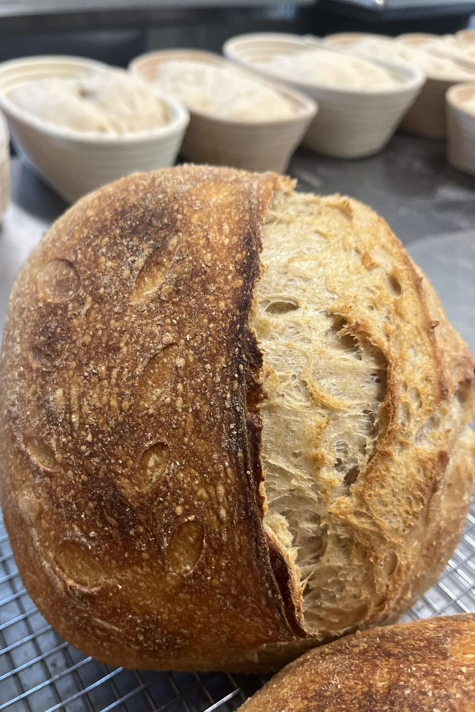 A round sourdough loaf, scored across the top and cut open, cooling on a wire rack in front of a row of proofing baskets.