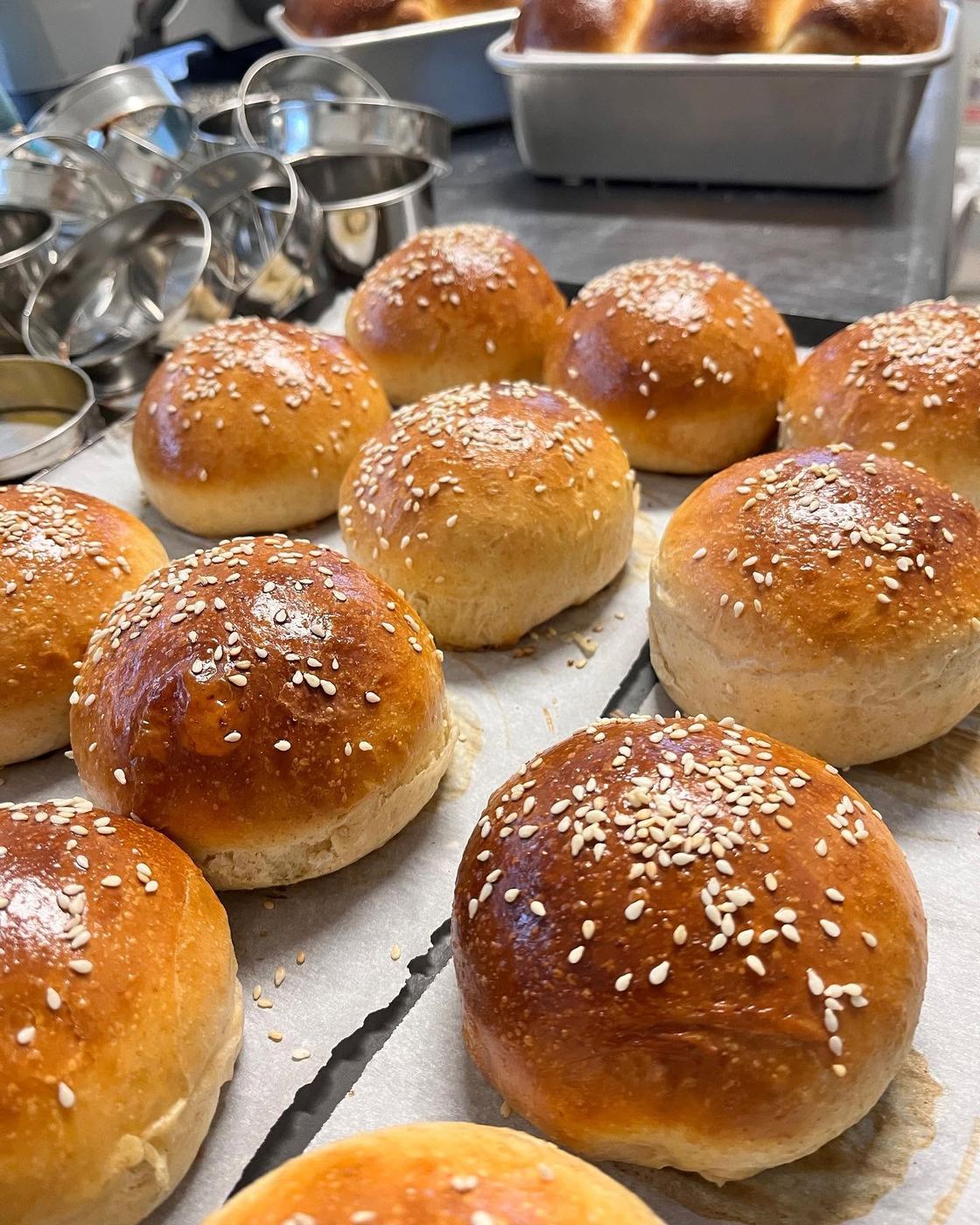A tray of glossy, sesame-topped rolls fresh from the oven, cooling beside stacked baking tins.